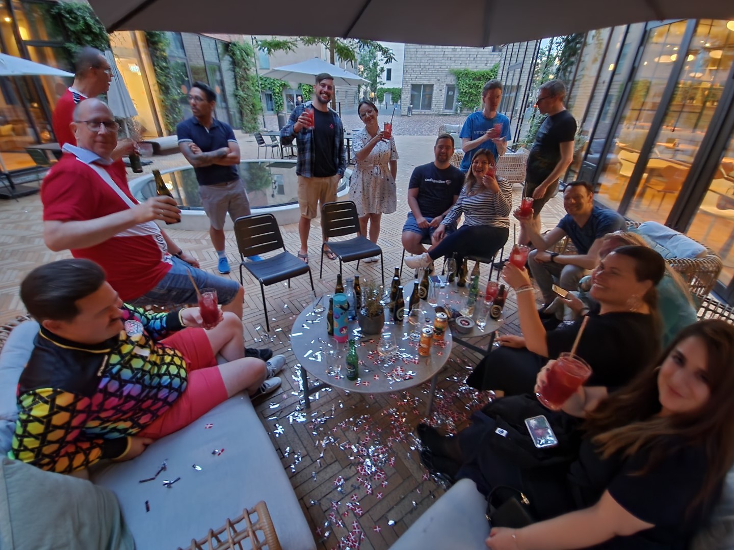 A group of people sitting at a small table outside the hotel with additional guests, confetti, and drinks.
